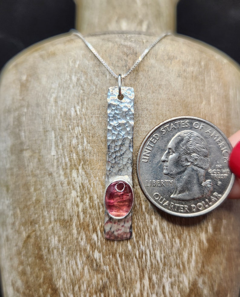 Puede incluir: Un collar con colgante de plata que presenta una barra de metal larga, rectangular y martillada con una piedra preciosa rosa ovalada engastada cerca de la base. Una moneda de 25 centavos de EE. UU. se coloca al lado para mostrar la escala.