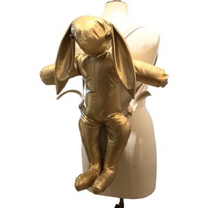 May include: A golden-colored bunny backpack with long ears and outstretched arms, attached to a mannequin. The bunny backpack is made of a shiny, soft material. The mannequin is a neutral color, and the background is white.