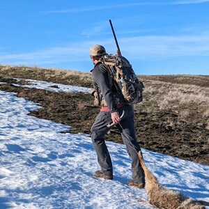 K&ouml;nnte beinhalten: Eine Person geht einen schneebedeckten Hang hinauf und tr&auml;gt ein Gewehr und einen gro&szlig;en Rucksack. Die Person zieht einen Kojoten hinter sich her. Der Himmel ist klar blau.