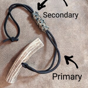 K&ouml;nnte beinhalten: Ein handgefertigter Geweihgriff mit einem schwarzen Kordelband. Die Kordel ist um das Geweih geknotet und hat einen geflochtenen Abschnitt mit gr&uuml;nen und beigen Akzenten. Die W&ouml;rter "Primary" und "Secondary" sind auf dem Bild geschrieben.