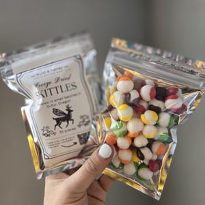 May include: Two clear, metallic pouches of freeze-dried Skittles. One pouch has a label with text and a deer graphic. The other pouch is filled with colorful, round candies. The candies have white, red, orange, yellow, green, and purple colors.