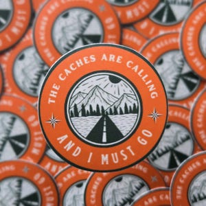 May include: A set of orange and white round stickers with a black and white illustration of a mountain road and the text "The Caches Are Calling And I Must Go".