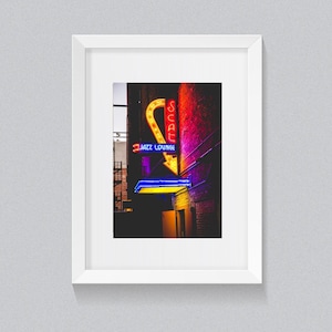 May include: Framed print of a neon sign for a jazz lounge. The sign features a yellow arrow, the word "SEAT" in red, and "JAZZ LOUNGE" in blue. The background is a brick wall.