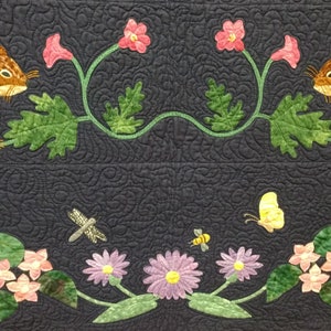 May include: A quilted wall hanging featuring two brown squirrels perched on a green vine with pink flowers. The bottom of the quilt features a green vine with yellow flowers, pink flowers, and a dragonfly.