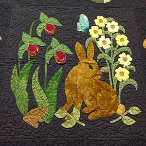 K&ouml;nnte beinhalten: Ein dunkelblauer gesteppter Stoff mit gestickten Bildern eines Waschb&auml;ren, eines Kaninchens, eines Kolibris und verschiedener Blumen. Der Stoff ist mit einem sich wiederholenden Muster aus Wirbeln verziert.