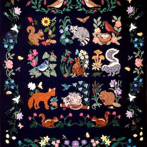 Puede incluir: Una colcha de fieltro azul oscuro con una variedad de animales y plantas bordadas. Los animales incluyen una ardilla, un mapache, un conejo, un zorro, un castor, una mofeta y p&aacute;jaros. Las plantas incluyen flores, hojas y hongos.