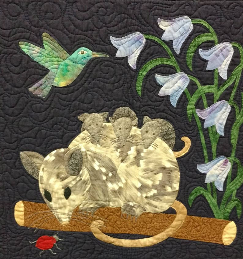 May include: A gray and white opossum with four babies on its back sits on a brown branch. A hummingbird with green and blue feathers flies above the opossum. The background is a dark blue fabric with a quilted pattern. There are several blue bell flowers with green stems and leaves.