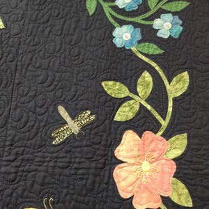 May include: A dark blue fabric with a vine design featuring pink, white, and blue flowers, green leaves, and yellow butterflies. The vine is quilted with a swirl pattern.