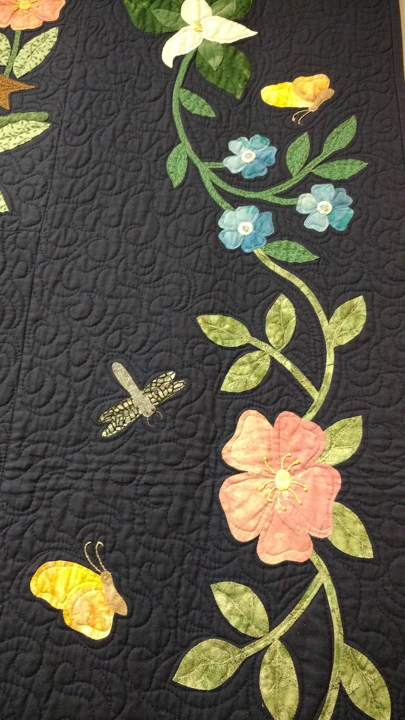 May include: A dark blue fabric with a vine design featuring pink, white, and blue flowers, green leaves, and yellow butterflies. The vine is quilted with a swirl pattern.