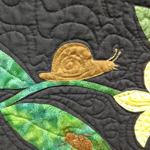 May include: A brown snail crawling on a green leaf with a brown beetle on another green leaf. The background is a dark blue fabric with a quilted pattern.