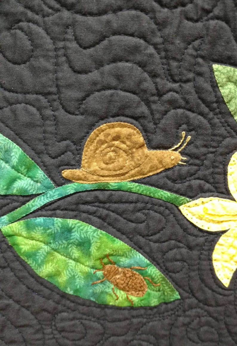 May include: A brown snail crawling on a green leaf with a brown beetle on another green leaf. The background is a dark blue fabric with a quilted pattern.