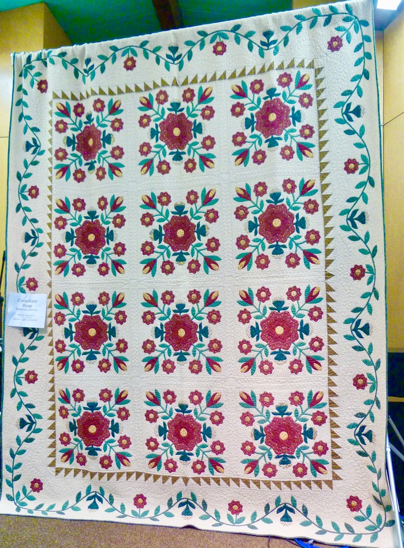 May include: A white quilt with a repeating pattern of red and green flowers. The quilt is edged with a green vine pattern. The quilt is titled "Calico Rose" and is displayed on a stand.