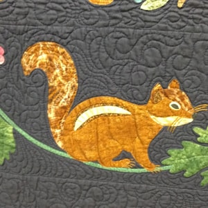 May include: A brown and white squirrel applique on a dark blue fabric background. The squirrel is surrounded by green leaves and pink flowers.