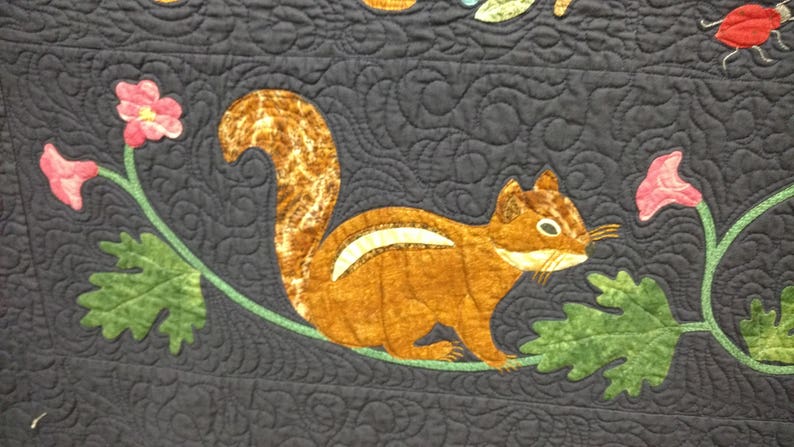 May include: A brown and white squirrel applique on a dark blue fabric background. The squirrel is surrounded by green leaves and pink flowers.
