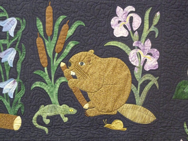 May include: A detailed quilt featuring a beaver holding a branch, surrounded by cattails, irises, and bluebell flowers. A lizard and snail are also depicted. The quilt has a dark blue background with quilted wavy lines.