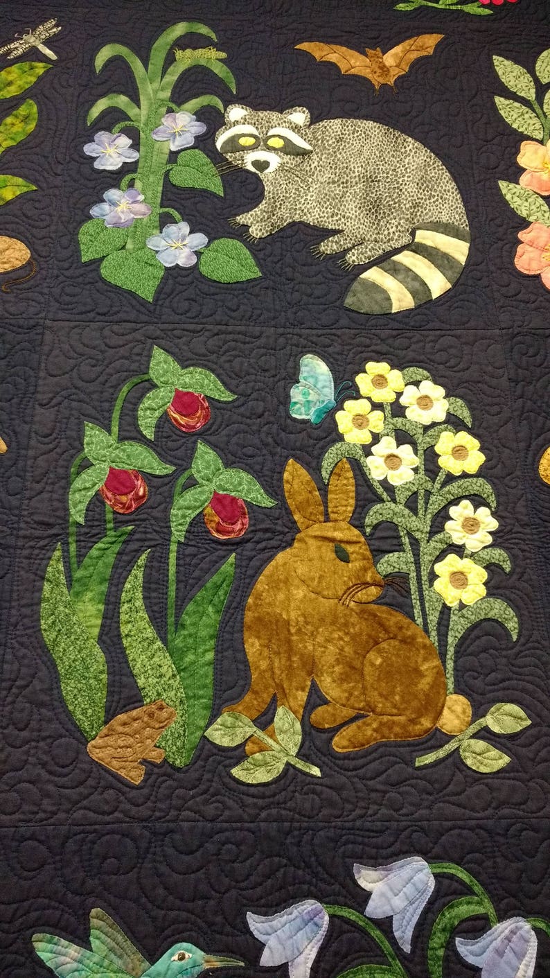 K&ouml;nnte beinhalten: Ein dunkelblauer gesteppter Stoff mit gestickten Bildern eines Waschb&auml;ren, eines Kaninchens, eines Kolibris und verschiedener Blumen. Der Stoff ist mit einem sich wiederholenden Muster aus Wirbeln verziert.