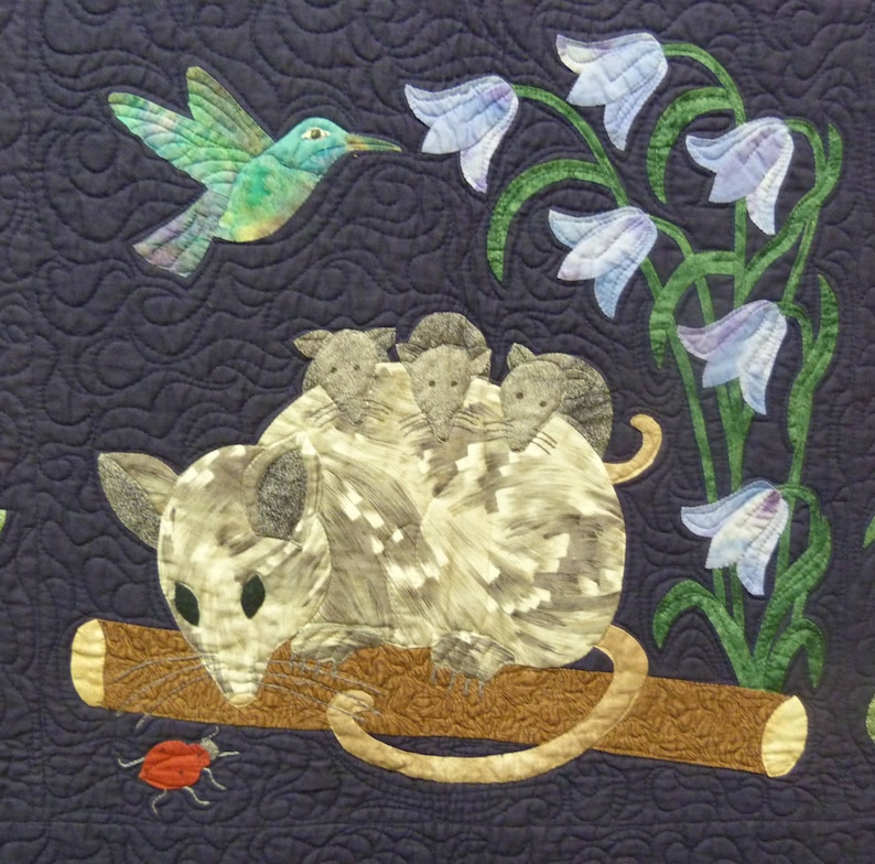 May include: A quilted wall hanging featuring a gray opossum with four babies on its back, perched on a brown branch. A hummingbird hovers above, and a red ladybug crawls on the branch. The background is a dark blue fabric with a swirling pattern.