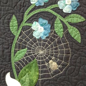 May include: A black fabric background with a white flower and green leaves.  There is a spider web with a spider in the center.  There are also blue flowers with white centers.