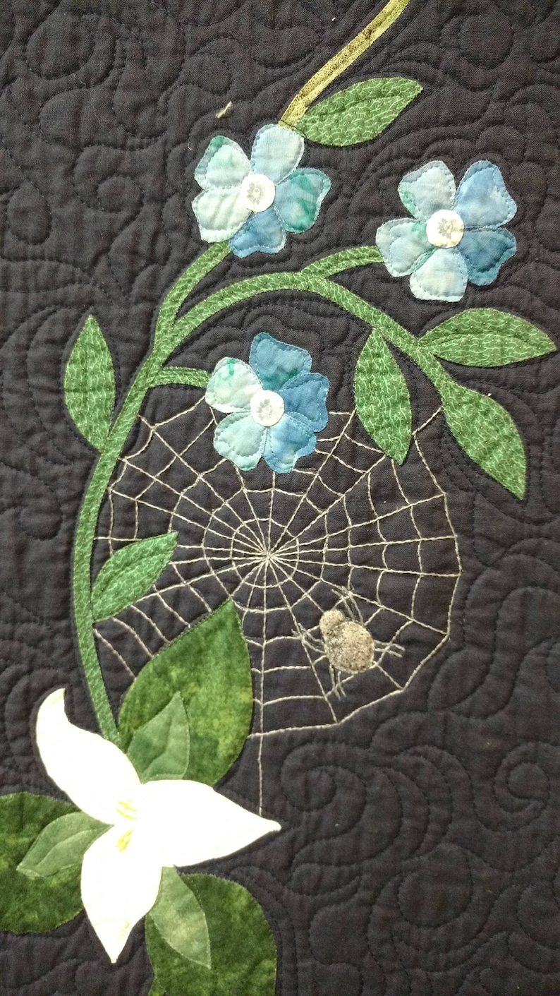 May include: A black fabric background with a white flower and green leaves.  There is a spider web with a spider in the center.  There are also blue flowers with white centers.