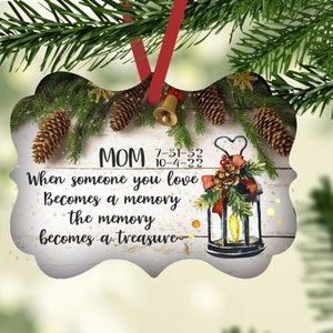 Puede incluir: Un adorno navideño de madera blanca con una linterna, piñas y vegetación. El adorno tiene el texto "MOM 7-31-32 10-4-22 When someone you love Becomes a memory the memory becomes a treasure."