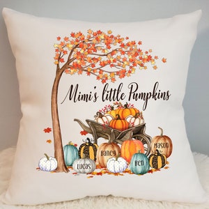 Pode incluir: Travesseiro branco com um design de outono. Uma árvore com folhas laranja e amarelas está no fundo. Uma carroça cheia de abóboras está em primeiro plano. O texto "Mimi's little Pumpkins" está escrito na almofada. As abóboras estão etiquetadas com os nomes Lucas, Bailey, Ben e Mason.
