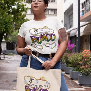 May include: White t-shirt and tote bag with the text "Forget the beads - throw me a Bunco" in purple, green, and gold. The tote bag is beige and the shirt is white. The design includes dice and a fleur-de-lis.