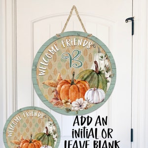 May include: Round wooden door hanger with a green and brown burlap background. The hanger features a watercolor illustration of pumpkins, gourds, and fall leaves. The text "Welcome Friends" is written in white on a green background. The text "Add an initial or leave blank" is written in black on a white background.
