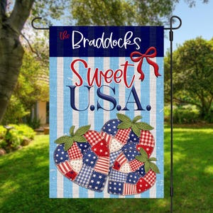 以下が含まれることがあります： 「Sweet U.S.A.」と「The Braddocks」の文字が入った愛国的なガーデンフラッグ。青と白のストライプの背景に、赤、白、青のパッチワークでハート型のイチゴのデザインが2つ描かれています。