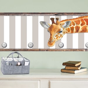 May include: A wooden framed wall decor with a white and gray striped background and a giraffe peeking over the top. The giraffe is brown and orange with black spots. There are five silver hooks on the frame.