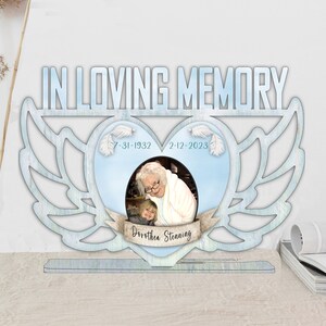 May include: A blue wooden memorial plaque with a heart-shaped cutout and angel wings. The heart has a photo of an older woman and a younger girl, with the dates "7-31-1932" and "2-12-2023" and the name "Dorothea Stenning" written below the photo.