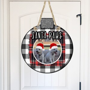May include: A round, black and white buffalo plaid Christmas ornament with a red plaid accent. The ornament has a photo of two white dogs wearing Santa hats. The text "Santa Paws" is written in black on the ornament.