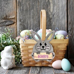 May include: A grey bunny rabbit with a sign that says "Bailey" is in a wicker basket filled with pastel coloured Easter eggs. The basket is sitting on a wooden surface.