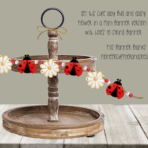 May include: A red and black ladybug and white daisy flower banner with holes for stringing. The banner is displayed on a tiered wooden tray. The text "Fits Banner Blanks at PioneerSupplierandcreations" is visible in the image.