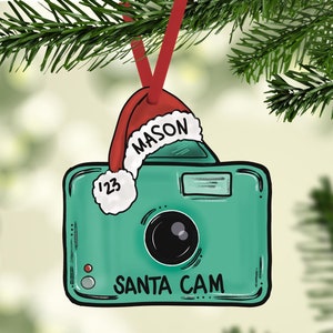 May include: A green camera ornament with a red Santa hat that says "MASON" on it. The camera has a white lens and says "SANTA CAM" on the bottom.