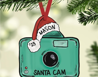 Könnte beinhalten: Ein grüner Kamera-Anhänger mit einer roten Weihnachtsmütze, auf der "MASON" steht. Die Kamera hat ein weißes Objektiv und unten steht "SANTA CAM".