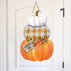 May include: A stacked pumpkin door hanger with a white pumpkin with gold polka dots, a yellow and white checkered pumpkin, and a large orange pumpkin with a "welcome fall" tag.