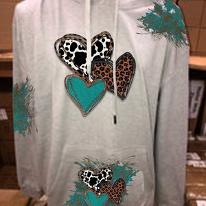 Könnte beinhalten: Ein hellgrauer Kapuzenpullover mit Kordelzug und Kängurutasche. Auf der Vorderseite befindet sich ein Design aus mehreren Herzen mit Kuhfellmuster, Leopardenmuster und türkisfarbenen Akzenten. Türkisfarbene Farbspritzer sind vorhanden.