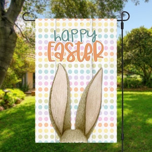 May include: A colorful garden flag with a white wooden background and a polka dot pattern. The text "Happy Easter" is written in orange and brown letters. Two bunny ears peek out from behind the wooden planks.