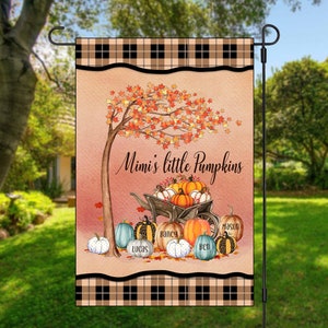 Könnte beinhalten: Eine Herbst-Gartenflagge mit kariertem Rand. Die Flagge zeigt eine skurrile Illustration eines Baumes mit orangefarbenen und roten Blättern, eine Schubkarre voller Kürbisse und den Text "Mimi's little Pumpkins". Die Kürbisse sind mit Namen beschriftet: Lucas, Bailey, Ben und Mason.