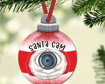 Könnte beinhalten: Roter und weißer Weihnachtsbaumschmuck mit Glitzer. Der Schmuck hat eine Kameraobjektiv mit zwei Händen darum und den Text "Santa Cam".