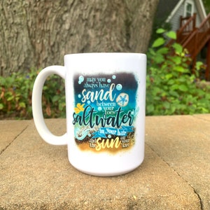 May include: White ceramic mug with a curved handle. The mug features the text "May you always have sand between your toes, saltwater in your hair, and the sun on your face" in blue, yellow, and white lettering, with a beach theme.