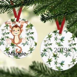 May include: Two round ceramic Christmas ornaments with a white background and green leafy patterns. One ornament features a cartoon monkey hanging from a branch with the name "Maggie" written below. The other ornament has the year "2019" written in the center.