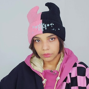 May include: A person wearing a black and pink beanie with a white eye and star design. The beanie is split down the middle, with one side pink and the other black. The person is also wearing a black and pink checkered jacket.
