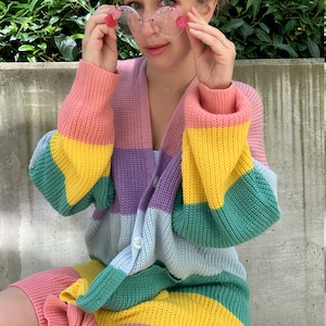 May include: A woman wearing a rainbow striped cardigan with a pink and yellow headband and round pink-tinted glasses.