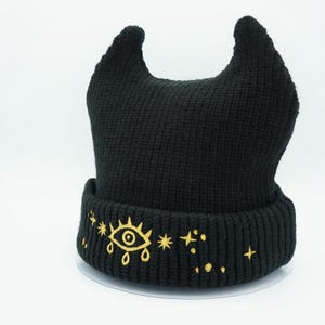 May include: Black knit beanie with a gold eye and star design. The beanie has two horns on top.