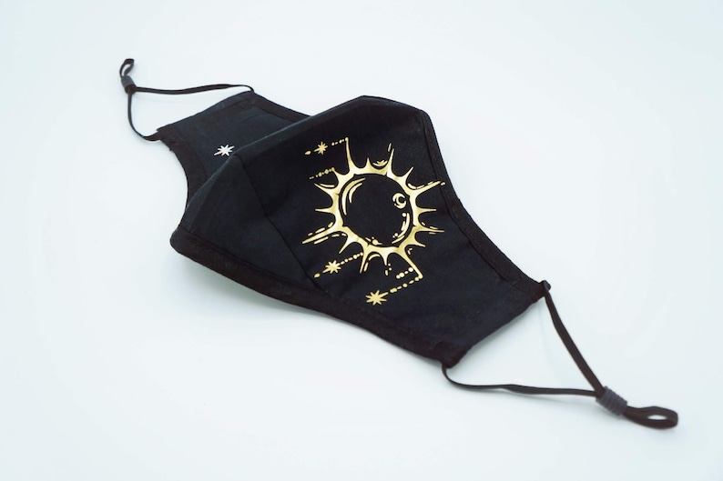 May include: Black face mask with a gold sun and moon design. The mask has adjustable ear loops.