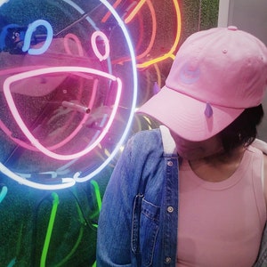 May include: A person wearing a pink denim jacket, a pink tank top, and a pink baseball cap with a blue crescent moon design. The person is standing in front of a neon sign that is shaped like a smiling face.
