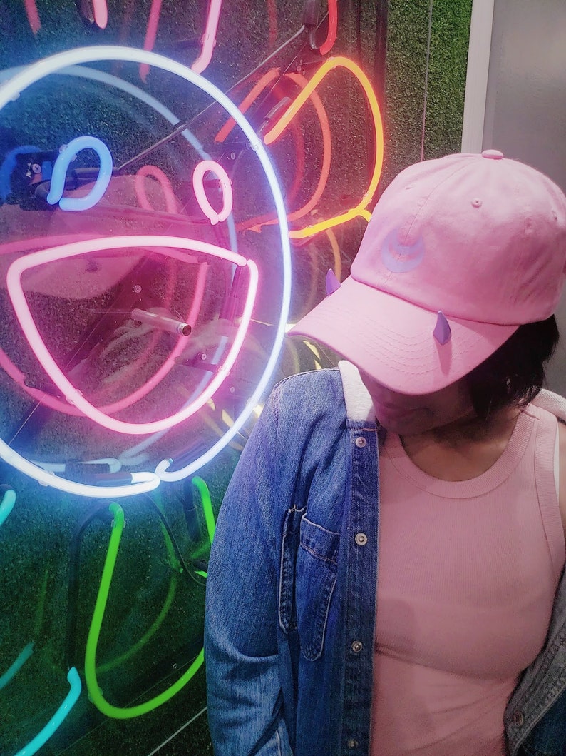 May include: A person wearing a pink denim jacket, a pink tank top, and a pink baseball cap with a blue crescent moon design. The person is standing in front of a neon sign that is shaped like a smiling face.