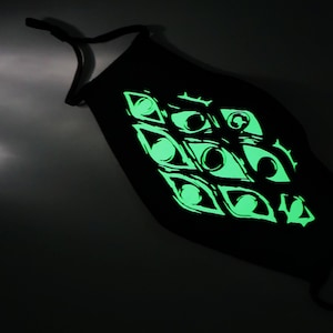 May include: Black face mask with a glowing green eye design. The mask has adjustable ear loops and a repeating pattern of stylized eyes. The design glows brightly against the dark fabric.
