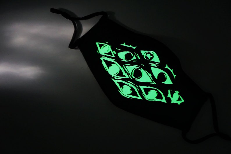 May include: Black face mask with a glowing green eye design. The mask has adjustable ear loops and a repeating pattern of stylized eyes. The design glows brightly against the dark fabric.